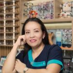 Smiling female optician in eyewear store standing behind counter surrounded by glasses displays.